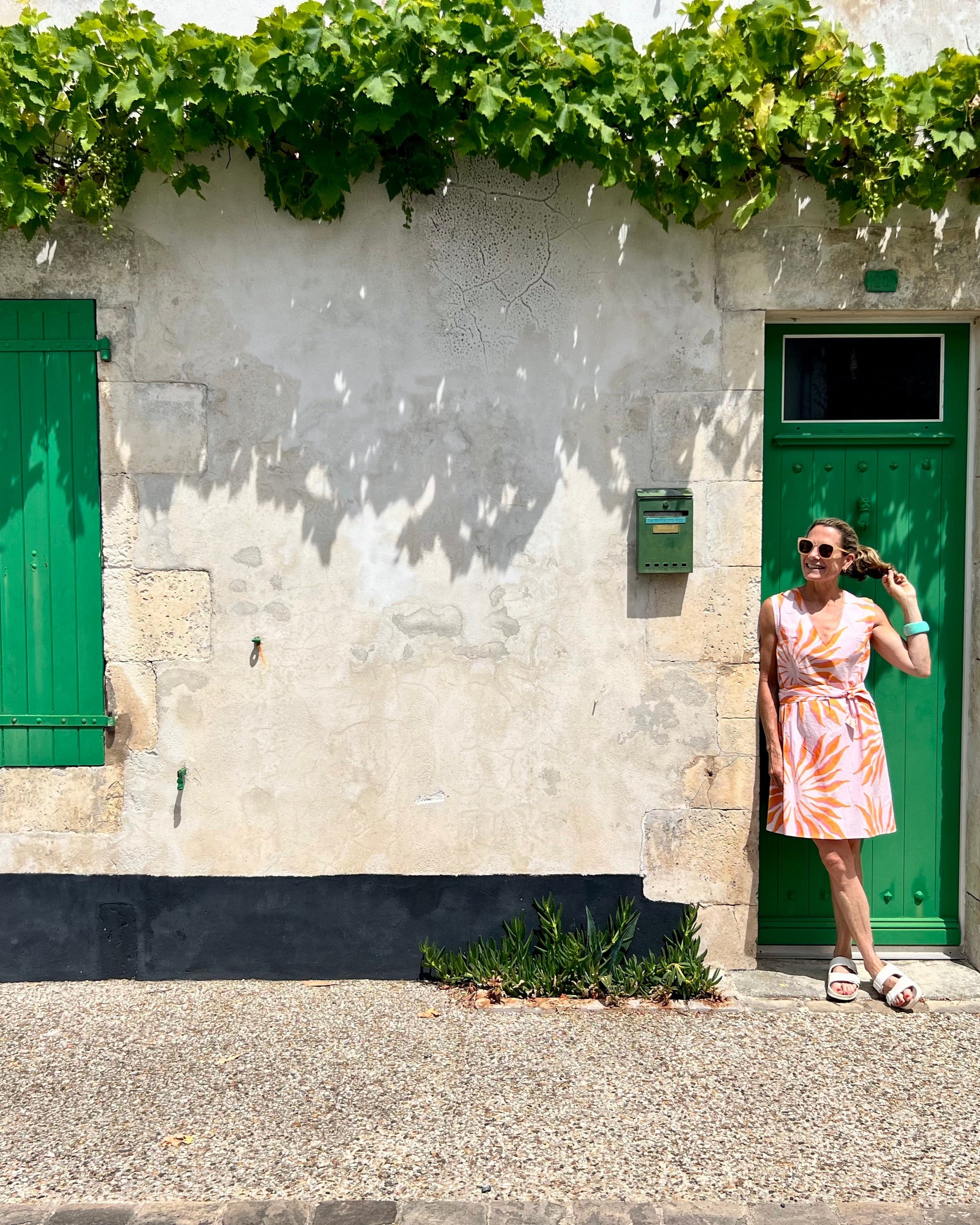 Woman in a pink and orange hand block-printed organic cotton dress standing in front of a white building with green doors and shutters.