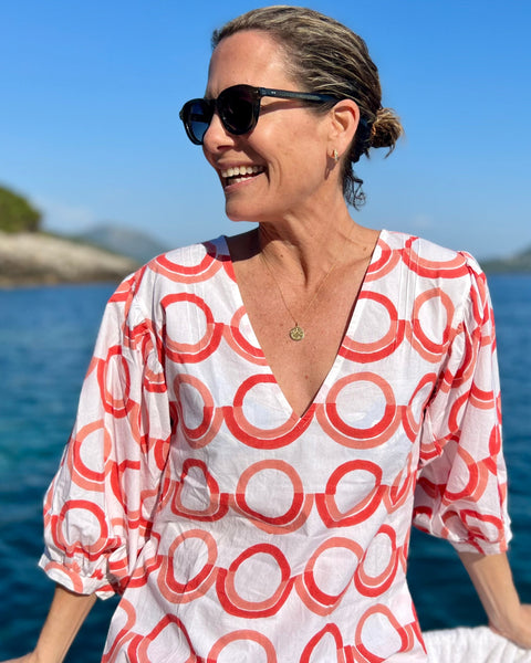 A person wearing a white blouse with a coral and pink geometric pattern, featuring a deep-V neck and puff sleeves, standing on a boat with a scenic background.