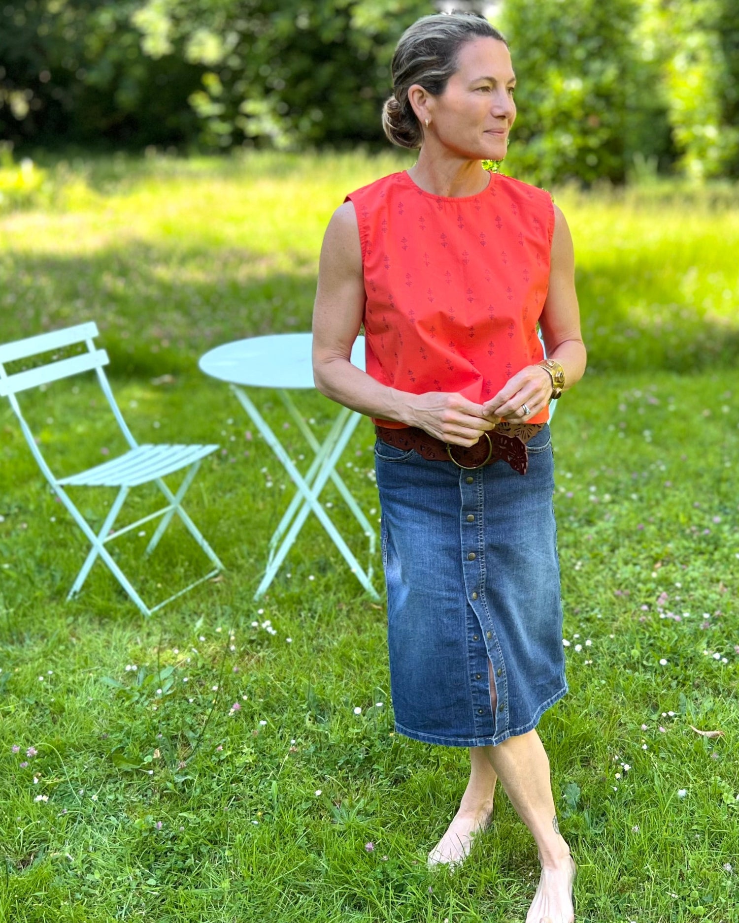 A person wearing a sleeveless, red and purple print, boxy top with a crew neck and a denim skirt in a grassy field