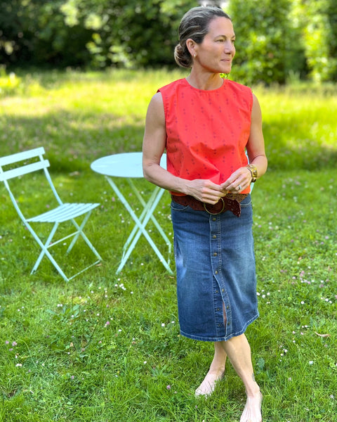 A person wearing a sleeveless, red and purple print, boxy top with a crew neck and a denim skirt in a grassy field