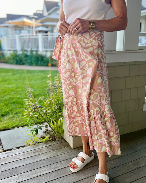 A woman standing on a porch with a blush pink and khaki animal print wrap skirt with a ruffle, paired with a white top and white birkenstocks