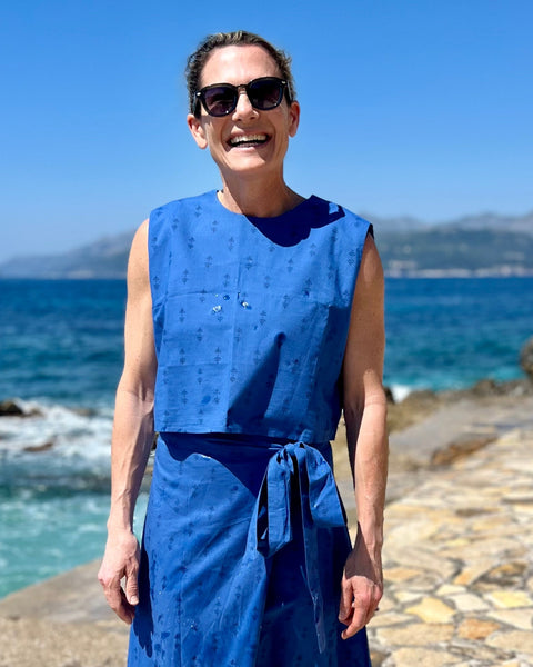 A person wearing a blue sleeveless crew neck crop top with a button closure at the neck, standing by the sea.