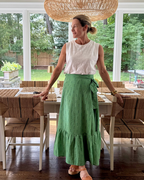 Woman in a white top and green hand block-printed maxi skirt standing in a dining room with wicker chairs and a large window.