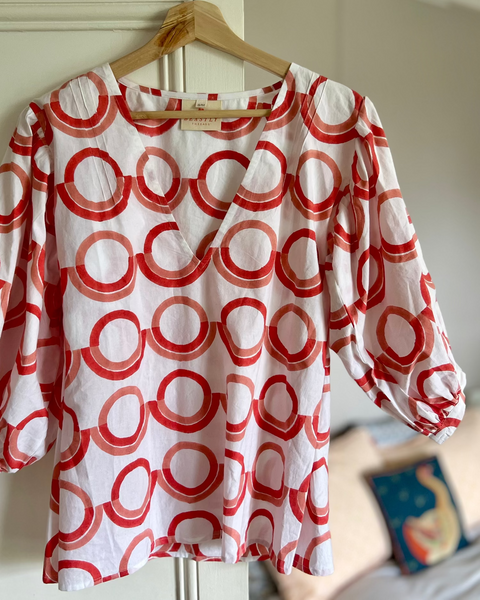A white blouse with a coral and pink geometric pattern, featuring a deep-V neck and puff sleeves on a hanger.