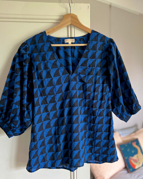 A navy blue and black blouse with a deep-V neck and 3/4 length puff sleeves, on a hanger in a bedroom.