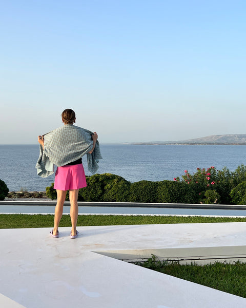 Woman in pink shorts wearing light blue cozy wrap/scarf with water in the background