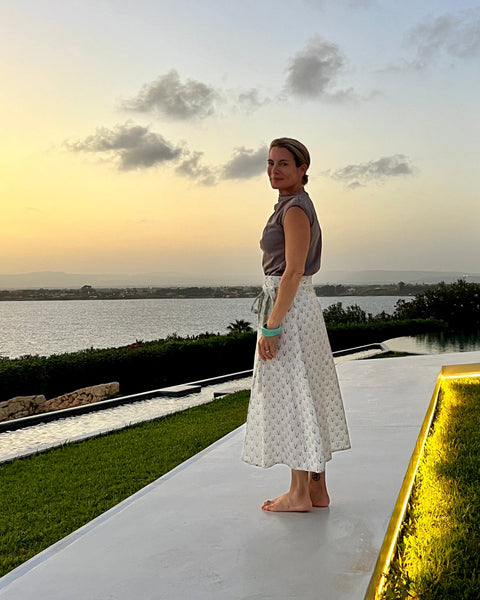 A person wearing an olive green, gray, and blue geometric patterned wrap skirt with a tea length cut and a taupe top at sunset