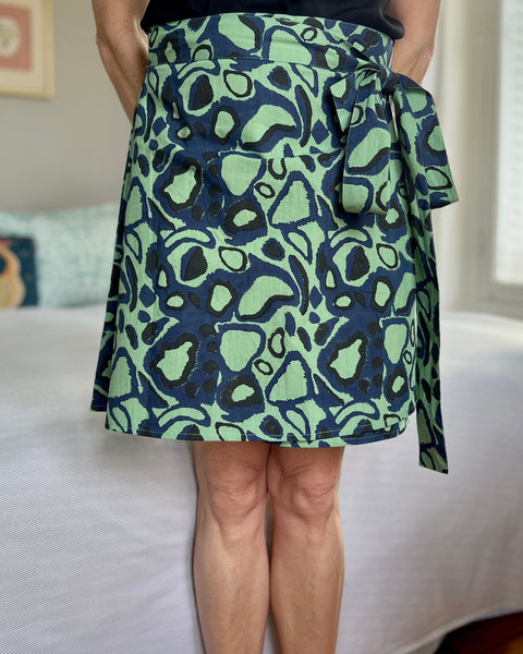 A person wearing a green wrap skirt with an animal print in navy blue, paired with a black top, standing in a room with natural lighting.
