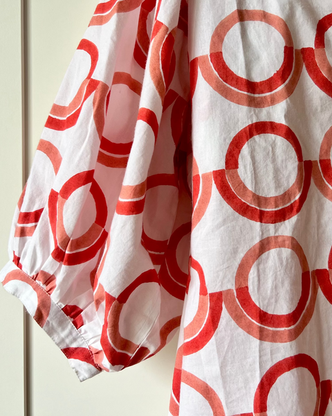 A white blouse with a coral and pink geometric pattern, featuring a deep-V neck and puff sleeves 