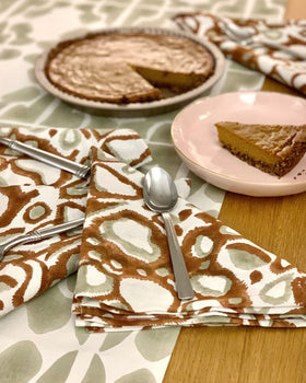 Table setting with rust wild dog hand block-printed napkins, silverware, and a slice of pie on a table runner