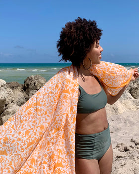 Woman on a beach holding an orange hand block-printed jaguar sarong / grand scarf with ocean and sunset in the background