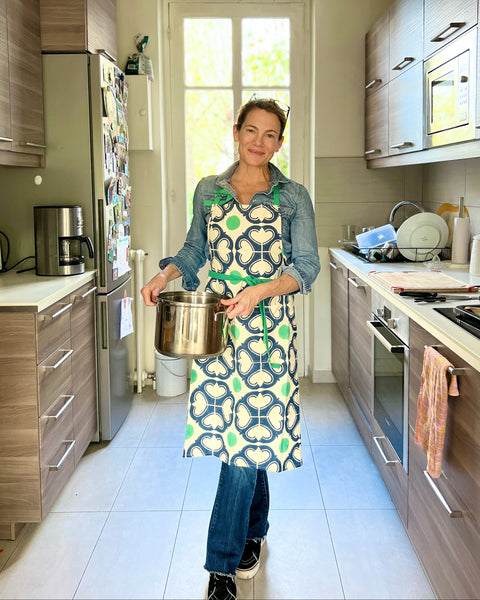 Woman wearing a navy blue, green, and off-white block-printed apron and jeans holding a pot in her kitchen