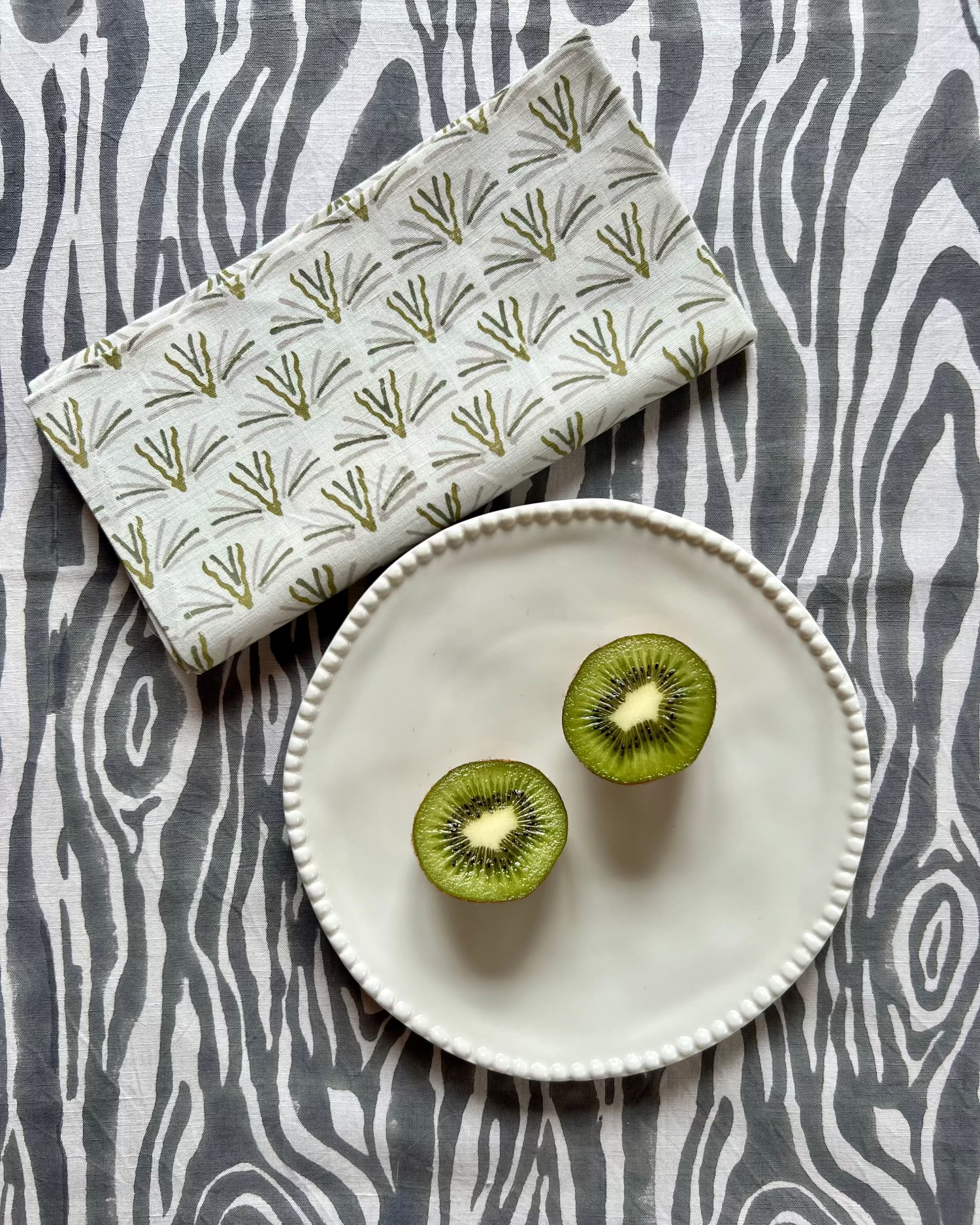 Two kiwi halves on a white plate on a gray and white faux bois hand block-printed organic cotton linen table runner with a blackbuck napkin