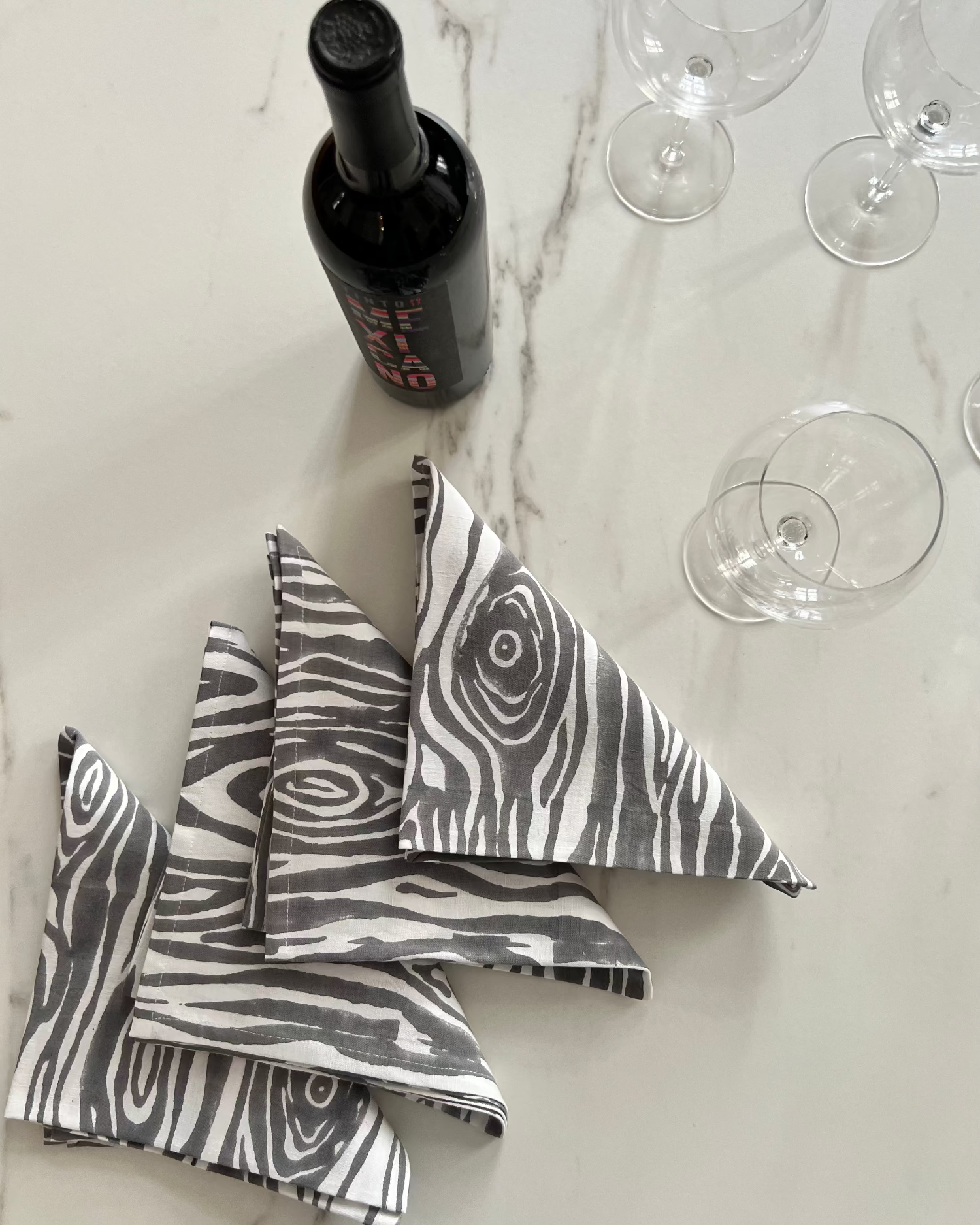 Gray and white faux bois print napkins on a marble surface with a bottle of wine and glasses.