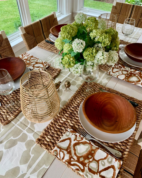 Table setting with straw placemats, hydrangeas, wooden bowls, and rust, khaki, and white hand block-printed napkin on a sand / khaki and white table runner