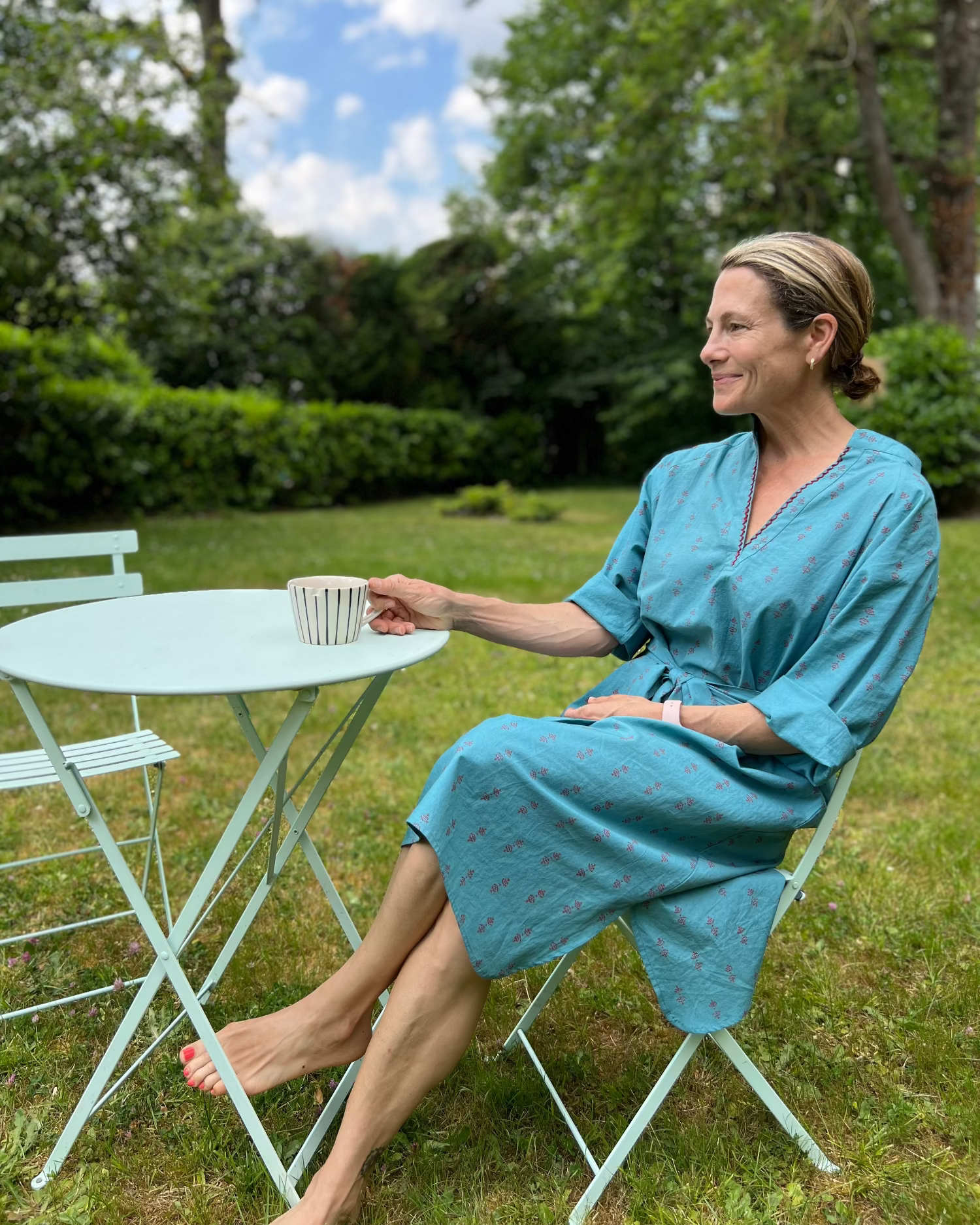 Woman in a teal midi dress sitting at a table outdoors, holding a cup.