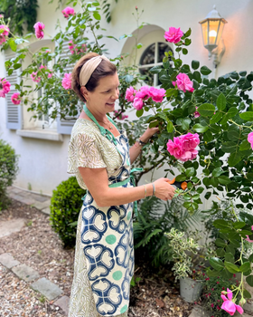 Woman wearing a navy blue, green, and off-white block-printed apron pruning roses in a french garden with a house in the background