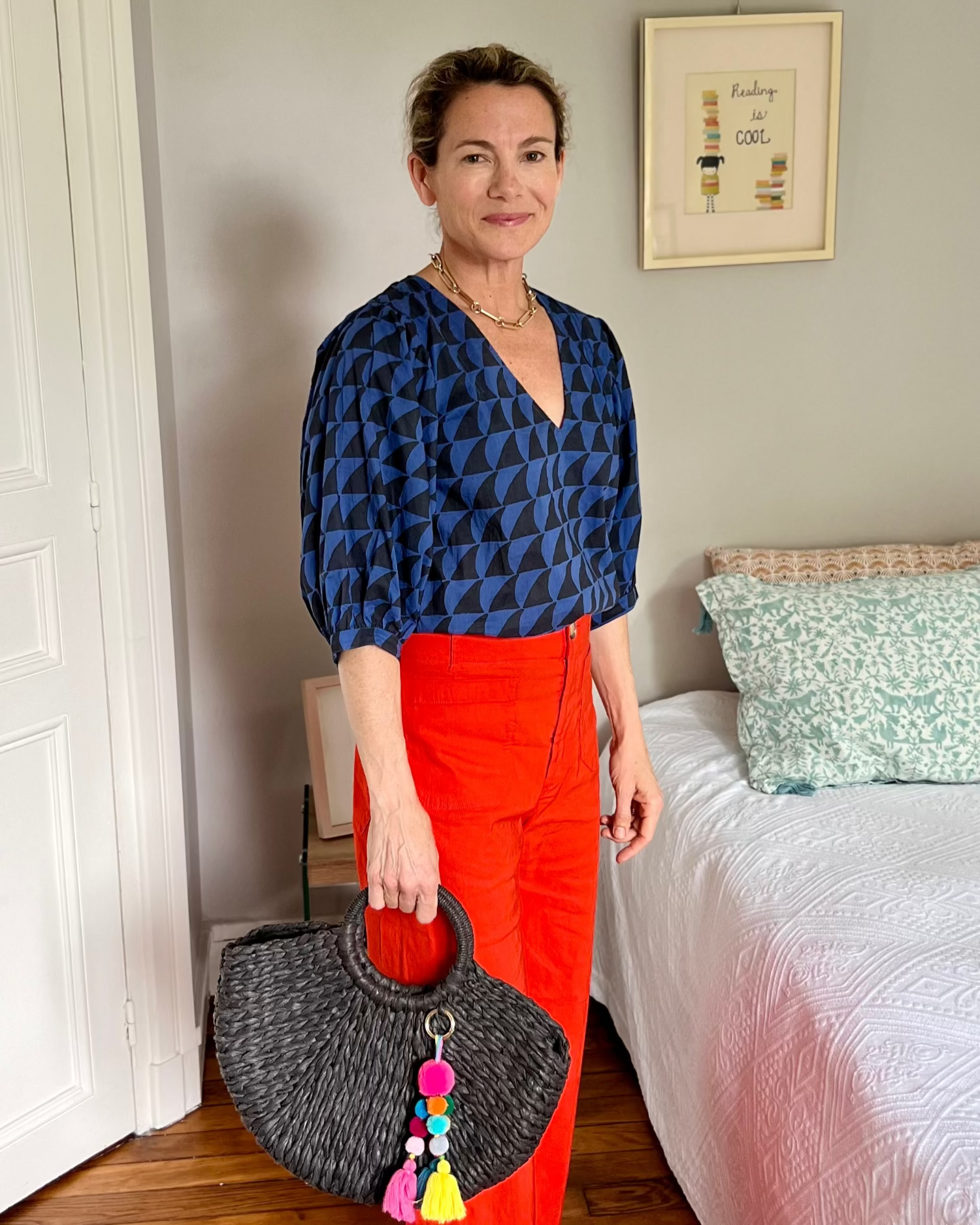 Woman in a blue and black shark patterned top and red pants holding a black handbag with colorful tassels in a bedroom.
