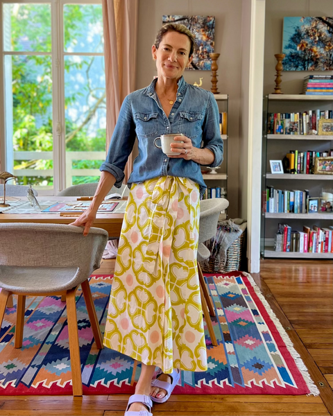 A person wearing a wrap skirt with a geometric pattern in yellow and pink on an off-white background and a denim shirt in a dining room