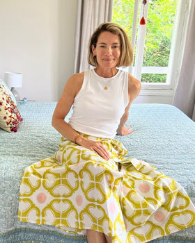A person seated on a bed wearing a wrap skirt with a geometric pattern in yellow and pink on an off-white background.