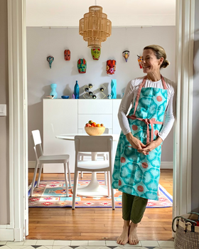 Woman wearing a turquoise and terracotta hand block-printed apron, standing in front of a dining area.