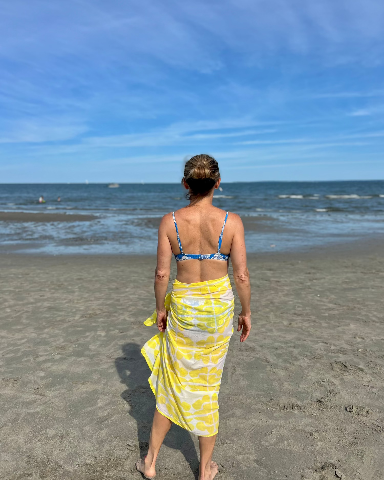 Woman walking on a beach wearing a yellow sarong and blue bikini top.