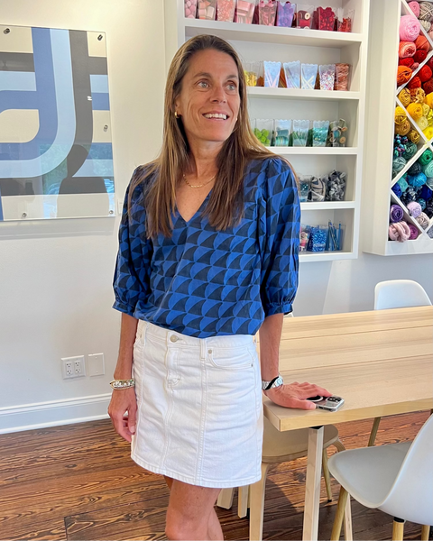 Woman in a blue and black shark print puff sleeve blouse standing in a room with shelves filled with colorful items, smiling.