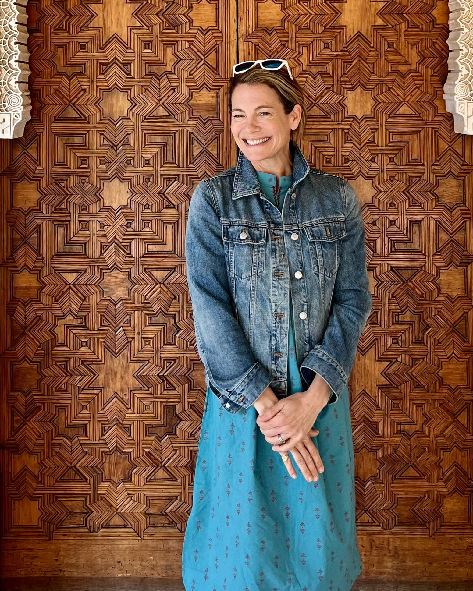 A woman wearing a teal midi dress with a red print and 3/4 length sleeves and a jean jacket in front of a Spanish door. The dress features an embroidered slash neckline and a long sash belt.