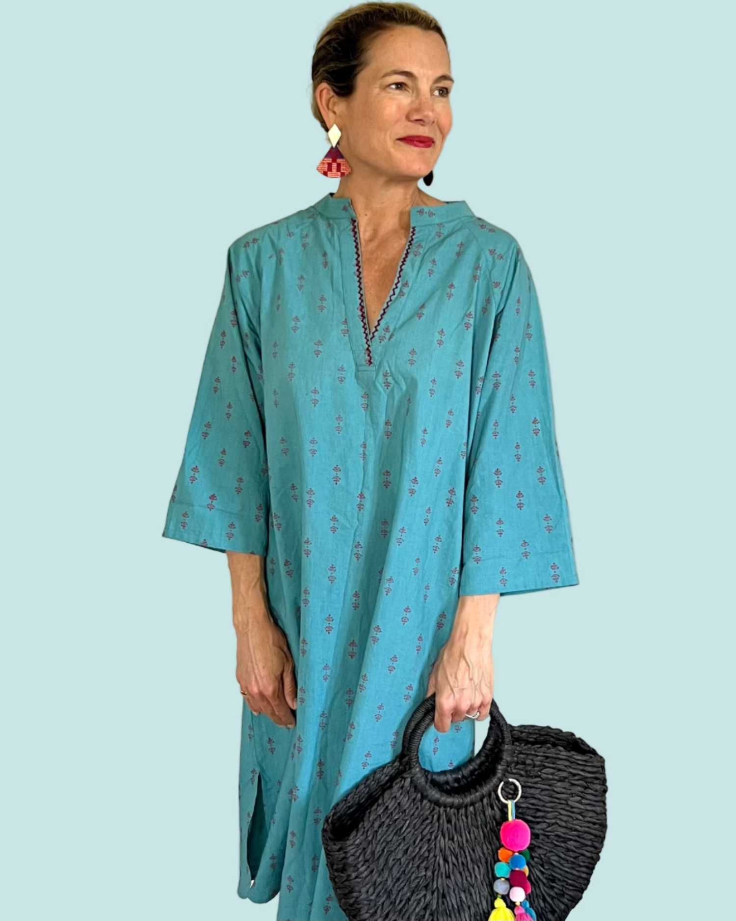 A woman wearing a teal midi dress with a red print and 3/4 length sleeves. The dress features an embroidered slash neckline and a long sash belt.