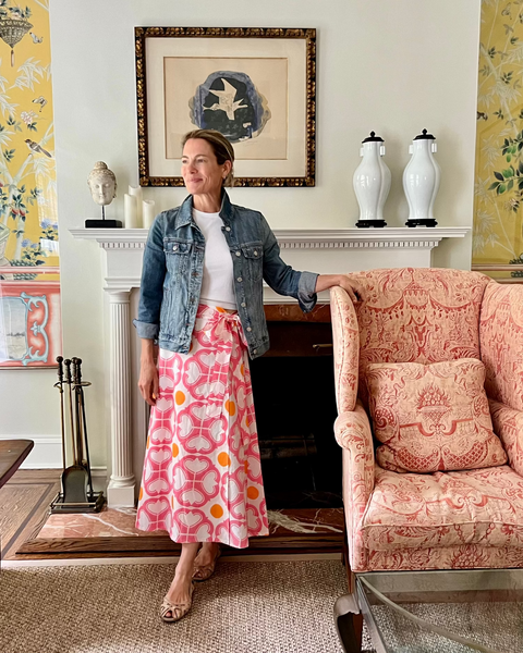 A woman wearing a watermelon pink and orange condor wrap skirt with a tea length cut, a white top, and a jean jacket in a beautiful sitting room