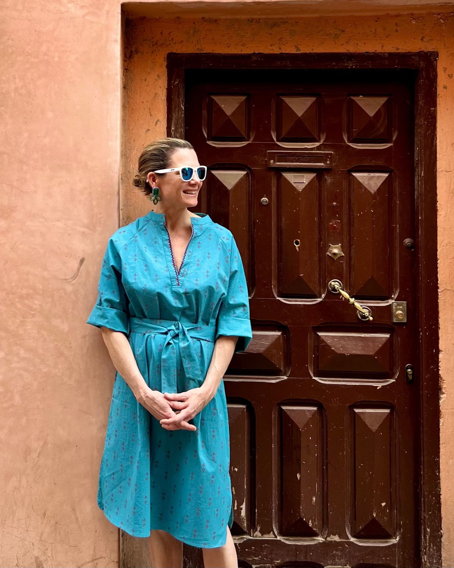 A woman wearing a teal midi dress with a red print and 3/4 length sleeves. The dress features an embroidered slash neckline and a long sash belt.