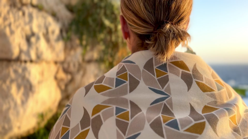 A video of a woman wearing a cream-colored cozy wrap/scarf with a geometric pattern featuring mustard yellow, taupe, and gray blue near the ocean