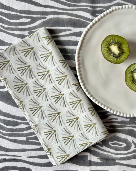 A folded cloth napkin with an olive green and gray pattern on a cream background, displayed on a dining table with a grey faux bois tablecloth and a kiwi on a plate.