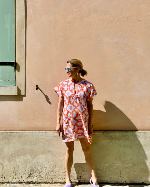 Woman in front of a wall wearing sunglasses and a coral, lavender, and white hand block-printed short kaftan dress 