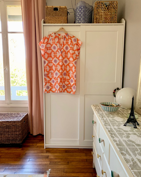 Coral, lavender, and white hand block-printed short kaftan dress hanging on an armoire in a sun-filled bedroom with a dresser and baskets