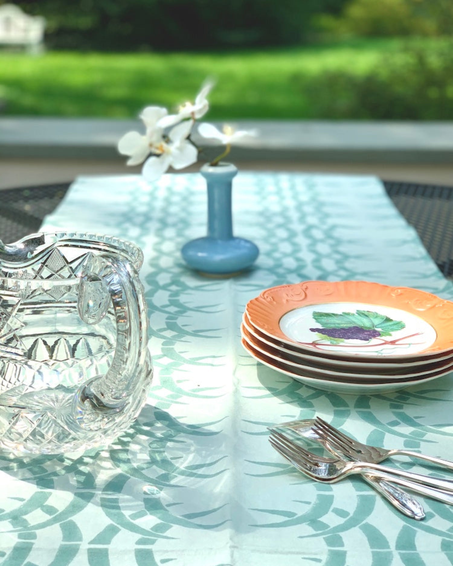 Table set with plates, flowers, utensils, and water pitcher on sage blue and ice blue rhino print table runner with green lawn in the background