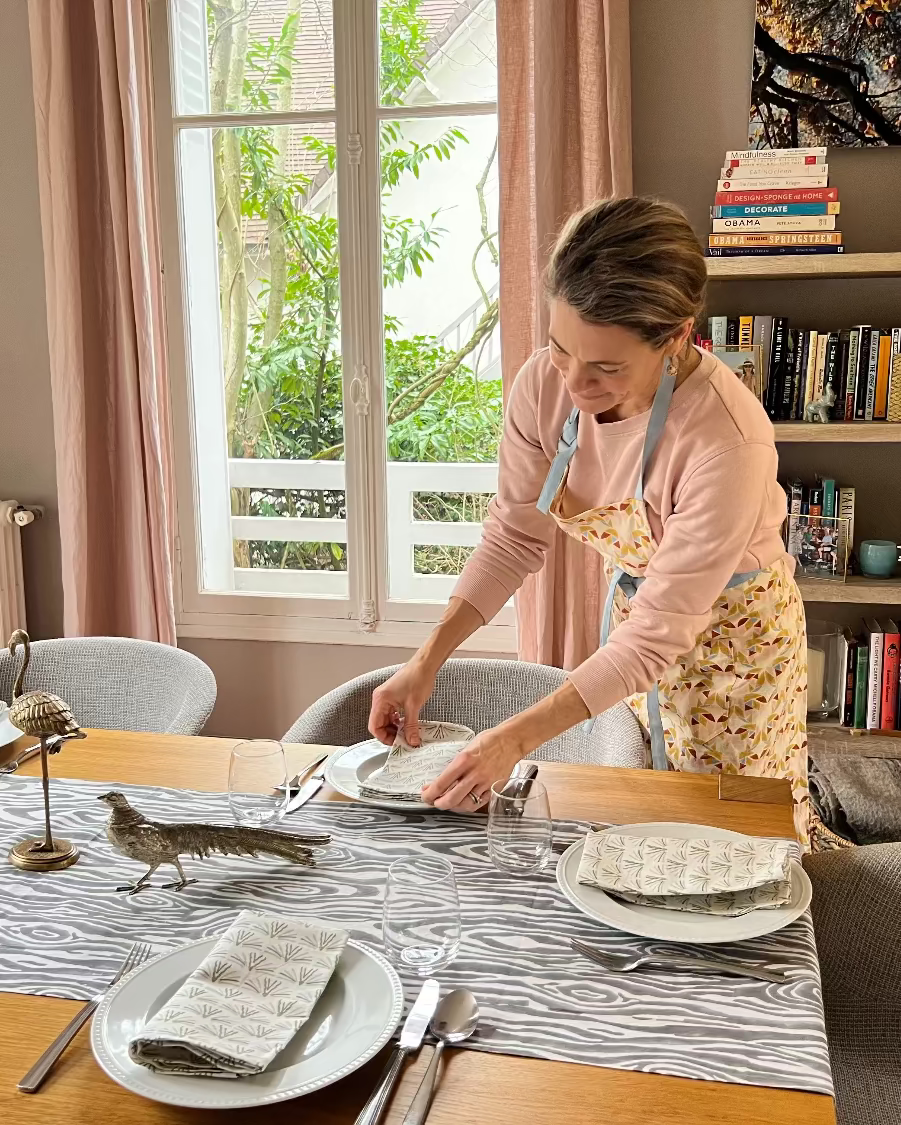 A woman setting a table with blackbuck organic cotton linen napkins, a gray and white faux bois hand block-printed organic cotton linen table runner, plates, utensils, and wine glasses.